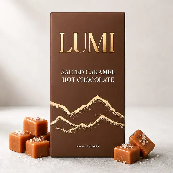 Box surrounded by caramels: “LUMI salted caramel hot chocolate box with gold mountain artwork, surrounded by caramel cubes and scattered salt.
