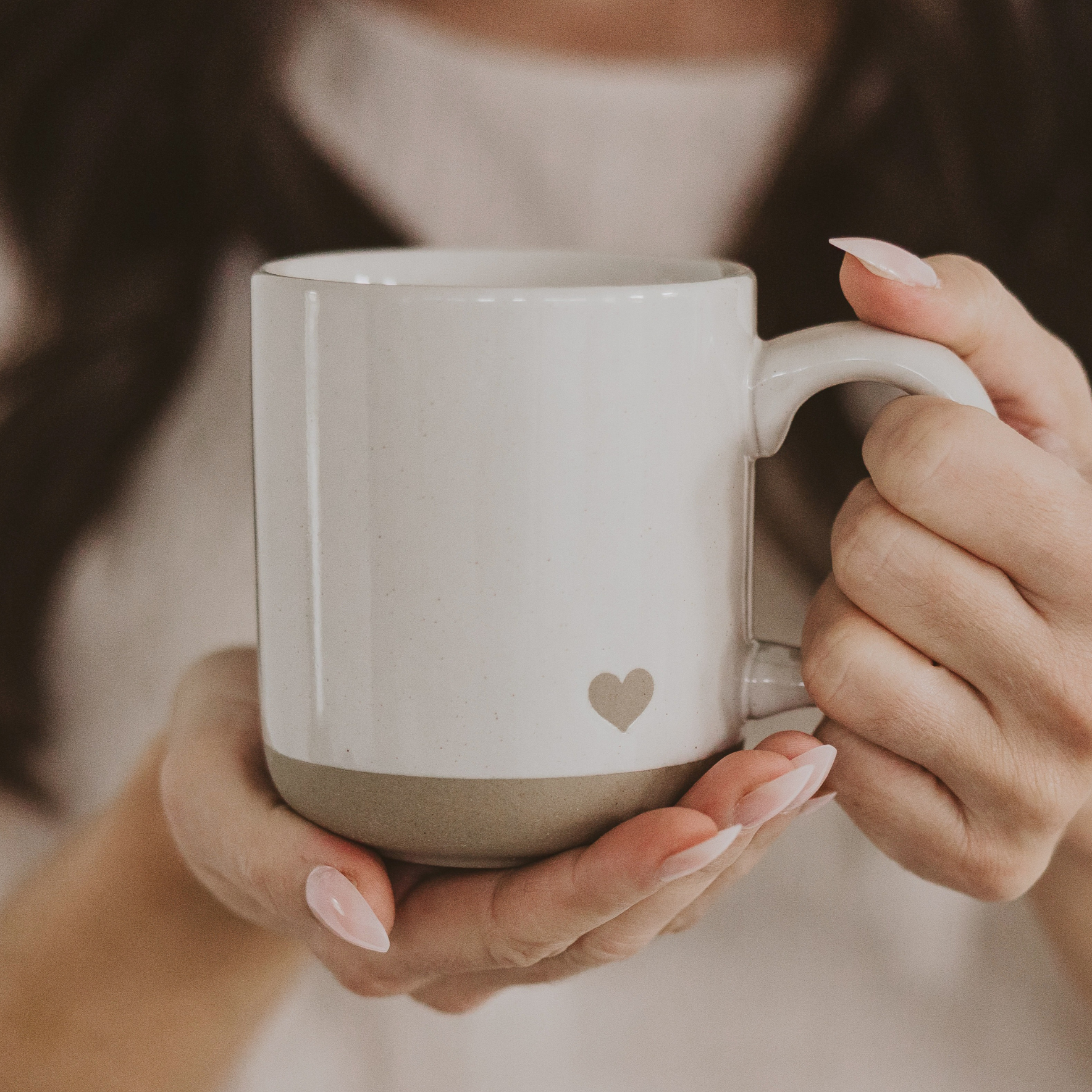 Person holding a white mug with a heart design, blurred background