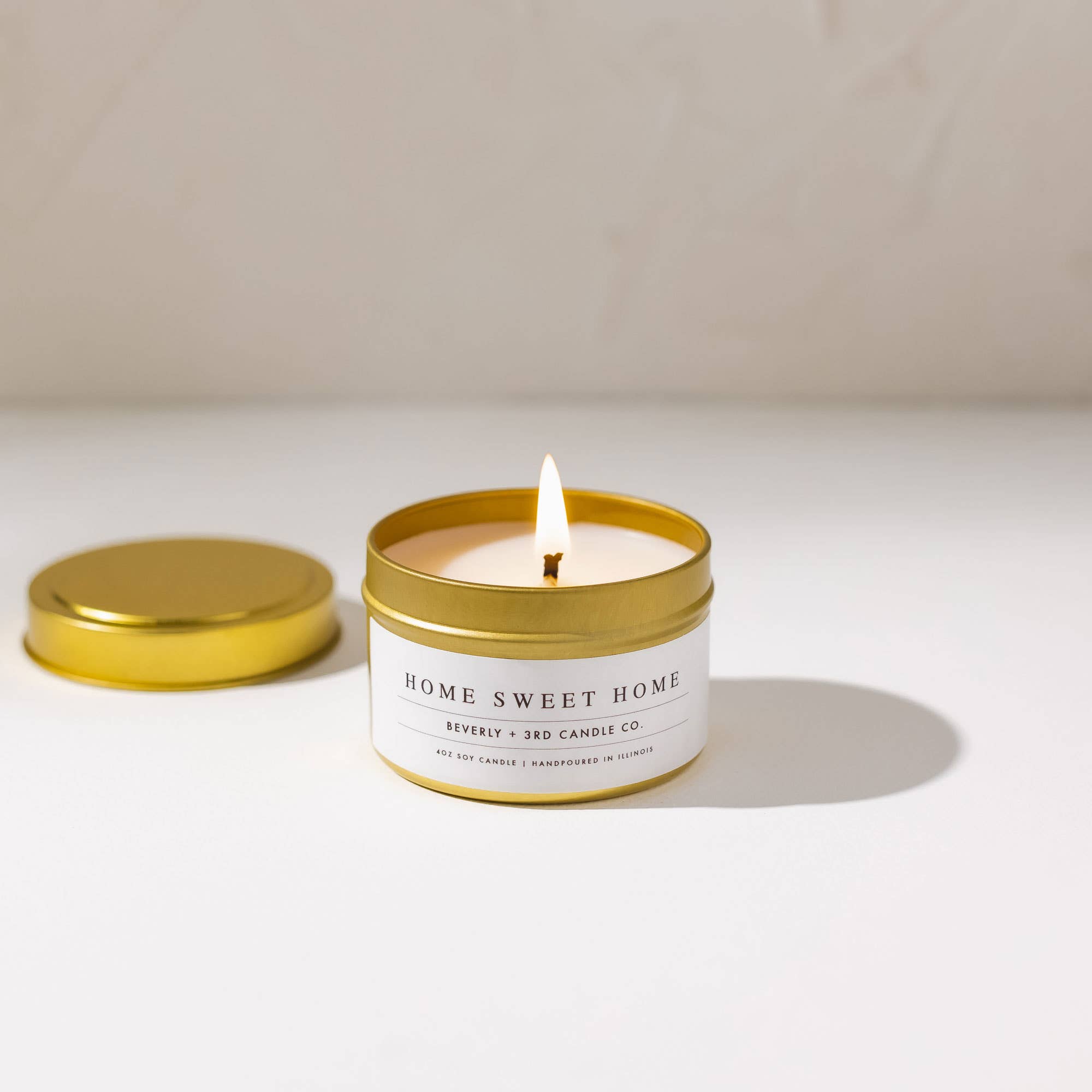 Candle in a gold tin labeled 'Home Sweet Home' on a white surface with a beige background