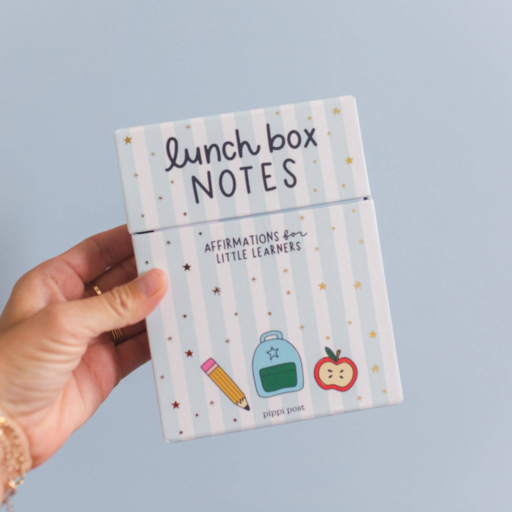 Hand holding a small notebook titled 'lunch box NOTES' with illustrations on a light blue background