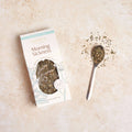 A package of Mummy's Organic Morning Sickness herbal tea sits on a light beige textured surface next to a small metal spoon filled with loose dried tea leaves.