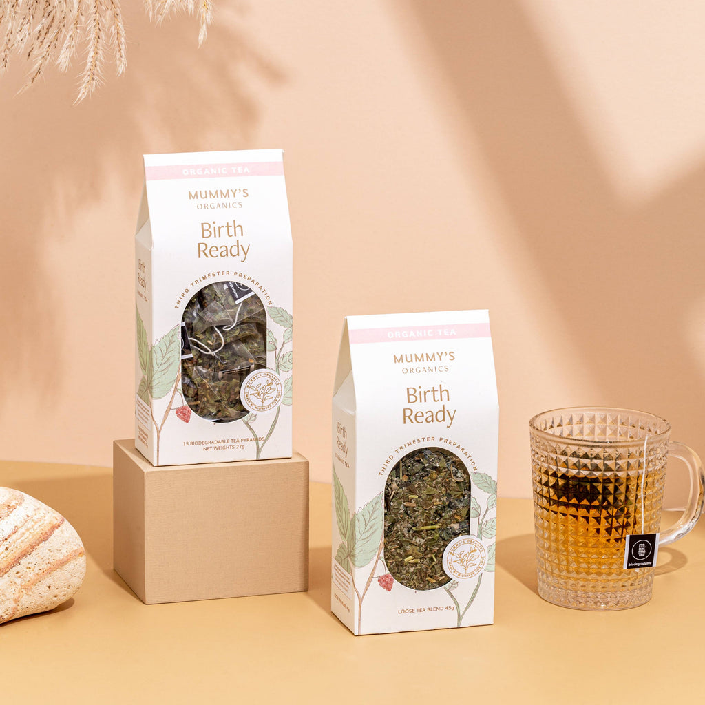 Two packages of Muminay's Organics Birth Ready Natural Tea displayed on a beige surface with a matching beige background, featuring a kraft box, a clear glass mug filled with tea, and dried pampas grass.