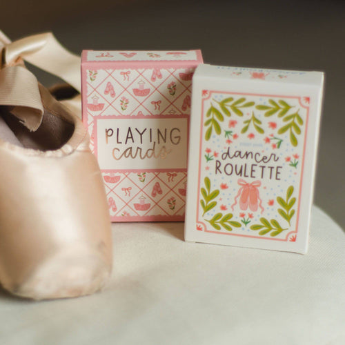A pair of pale pink satin pointe shoes and two decorative boxes of dancing cards on a soft white surface. One box reads “PLAYING cards” and the other reads “dancer ROULETTE,” both with pink and green ballet-themed illustrations.