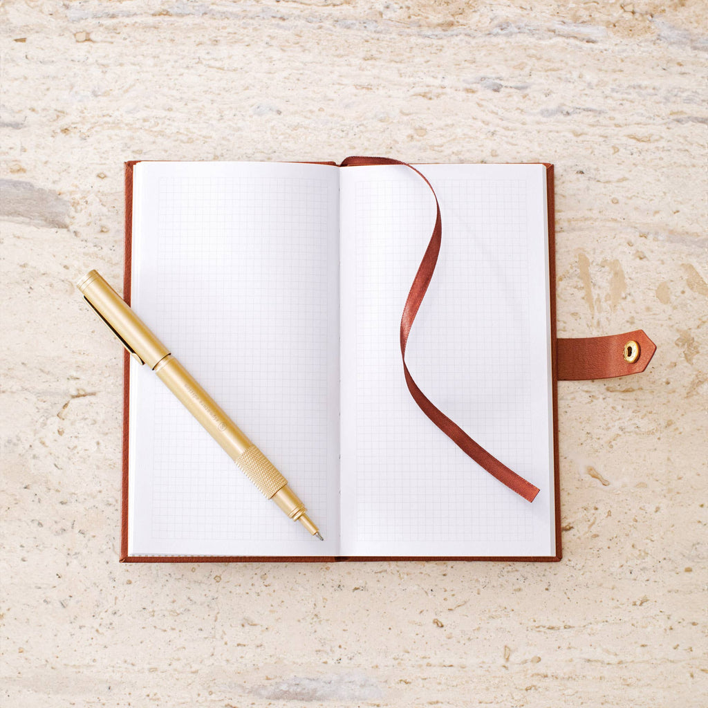 An open grid notebook with a brown leather cover and ribbon bookmark, styled with a gold pen on a light beige stone surface.