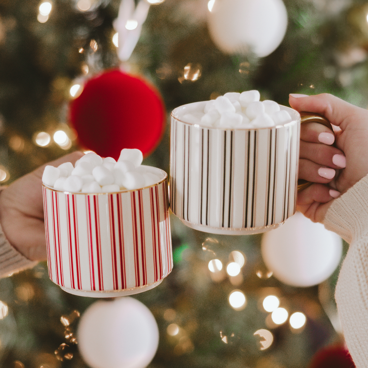 Red and green striped mugs with gold handles filled with marshmallows, held beside a decorated Christmas tree.

