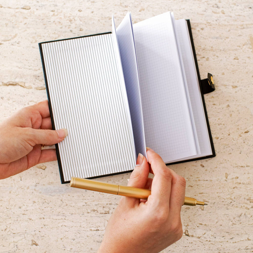 Open Signature Edition black faux leather pocket notebook showing lined pages, held in hand beside a gold pen. Compact notebook with a snap closure for journaling, note-taking, work, and everyday organization.