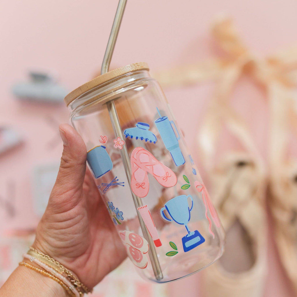 A hand holds a clear glass jar tumbler with a bamboo lid and metal straw. The transparent cup is decorated with pastel blue and pink illustrations of ballet flats, a bathtub, a trophy, and small flowers.