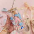 A hand holds a clear glass jar tumbler with a bamboo lid and metal straw. The transparent cup is decorated with pastel blue and pink illustrations of ballet flats, a bathtub, a trophy, and small flowers.