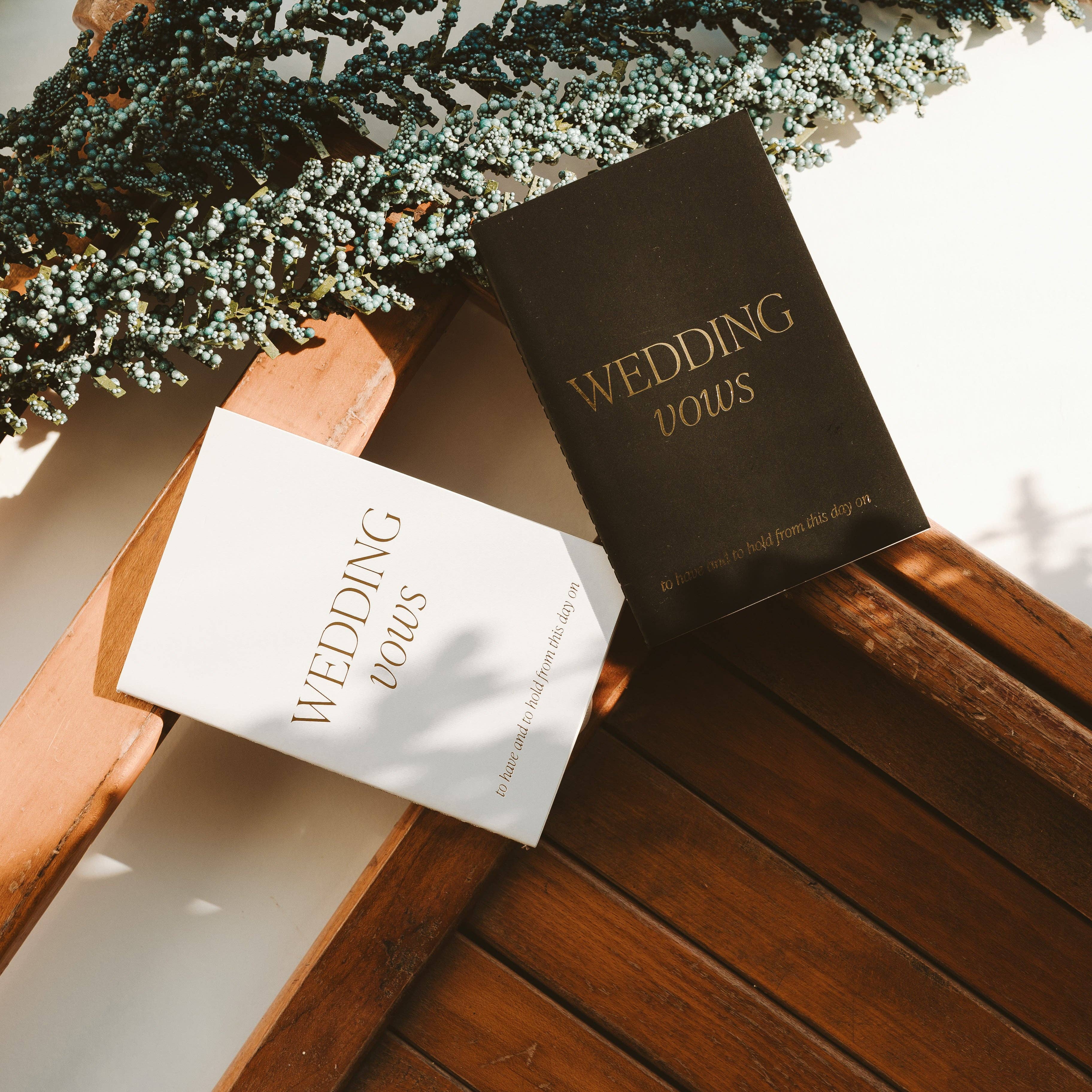 Two wedding vow books on a wooden surface with greenery in the background