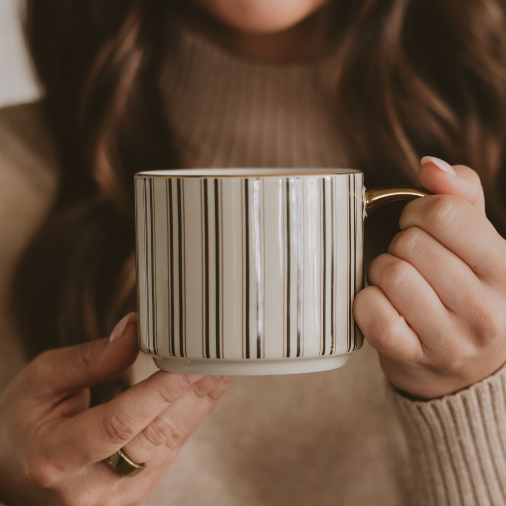 Person in a beige sweater holding a green stripe coffee mug with a gold handle and rim.
