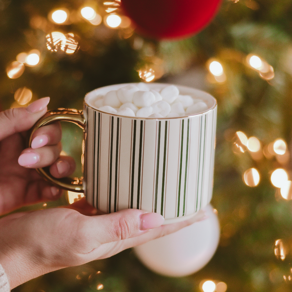 Green stripe mug with a gold handle filled with marshmallows, held in front of a glowing Christmas tree.
