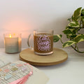 Glass mug with 'Eat Sleep Read' text on a wooden coaster next to a candle and book.