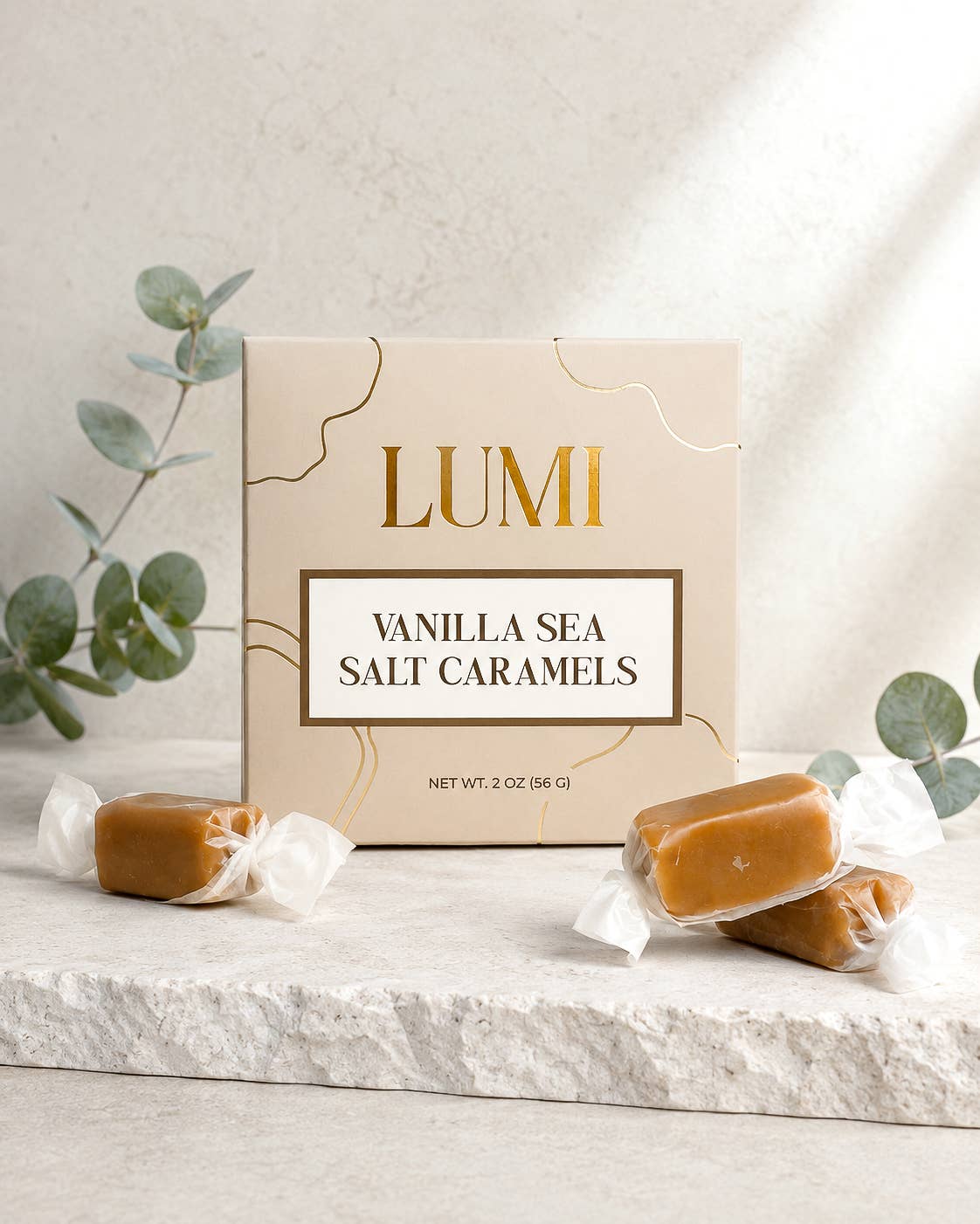 LUMI Vanilla Sea Salt Caramels in a beige gift box with gold accents, styled on a white stone surface with wrapped caramel bars and eucalyptus branches.