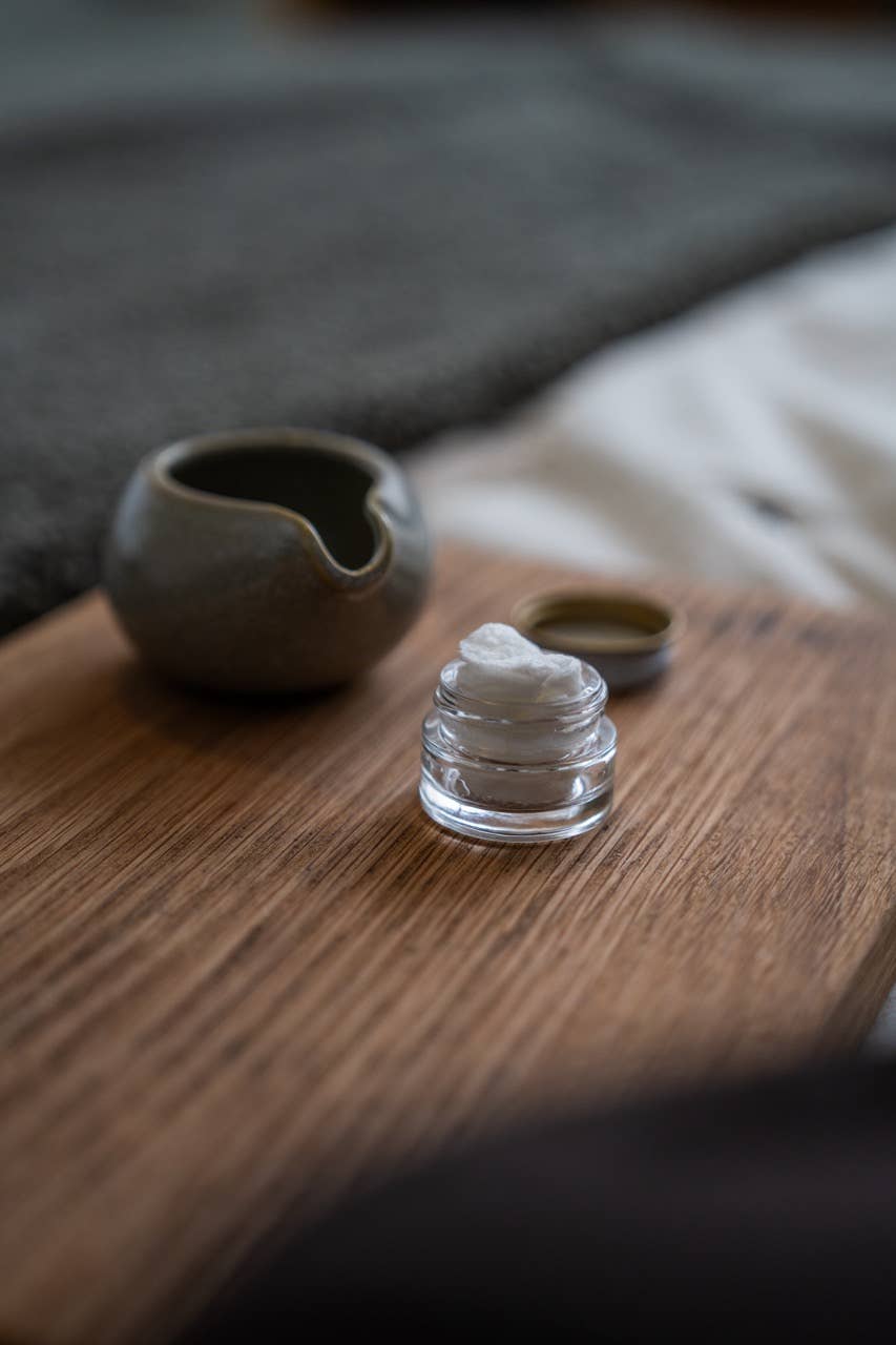 Small glass container with a white substance on a wooden surface next to a ceramic cup.