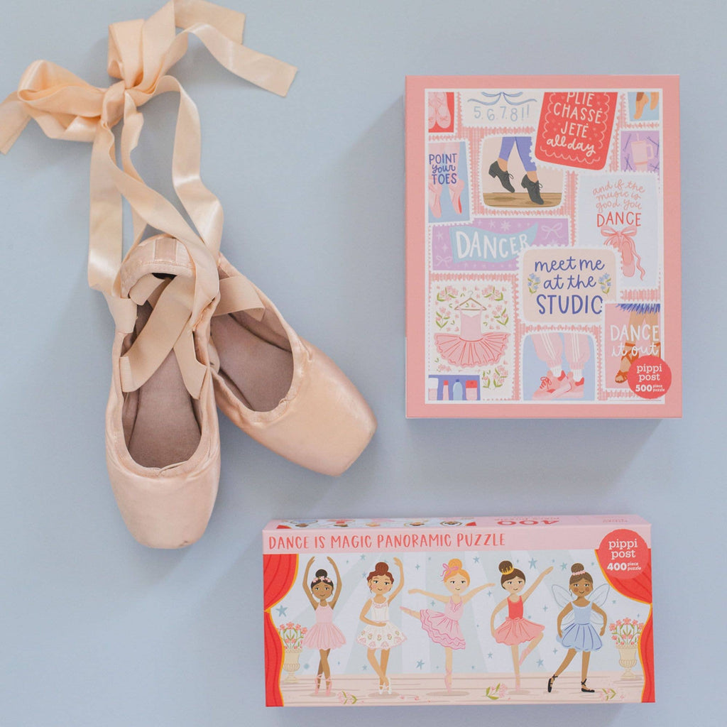 A pair of pale pink satin pointe shoes with long tied ribbons, shown beside two product boxes on a light blue background. The top box is pink with colorful ballet-themed illustrations and text, and the bottom box is red with ballerina artwork and the title 'Dance is Magic Panoramic Puzzle'.
