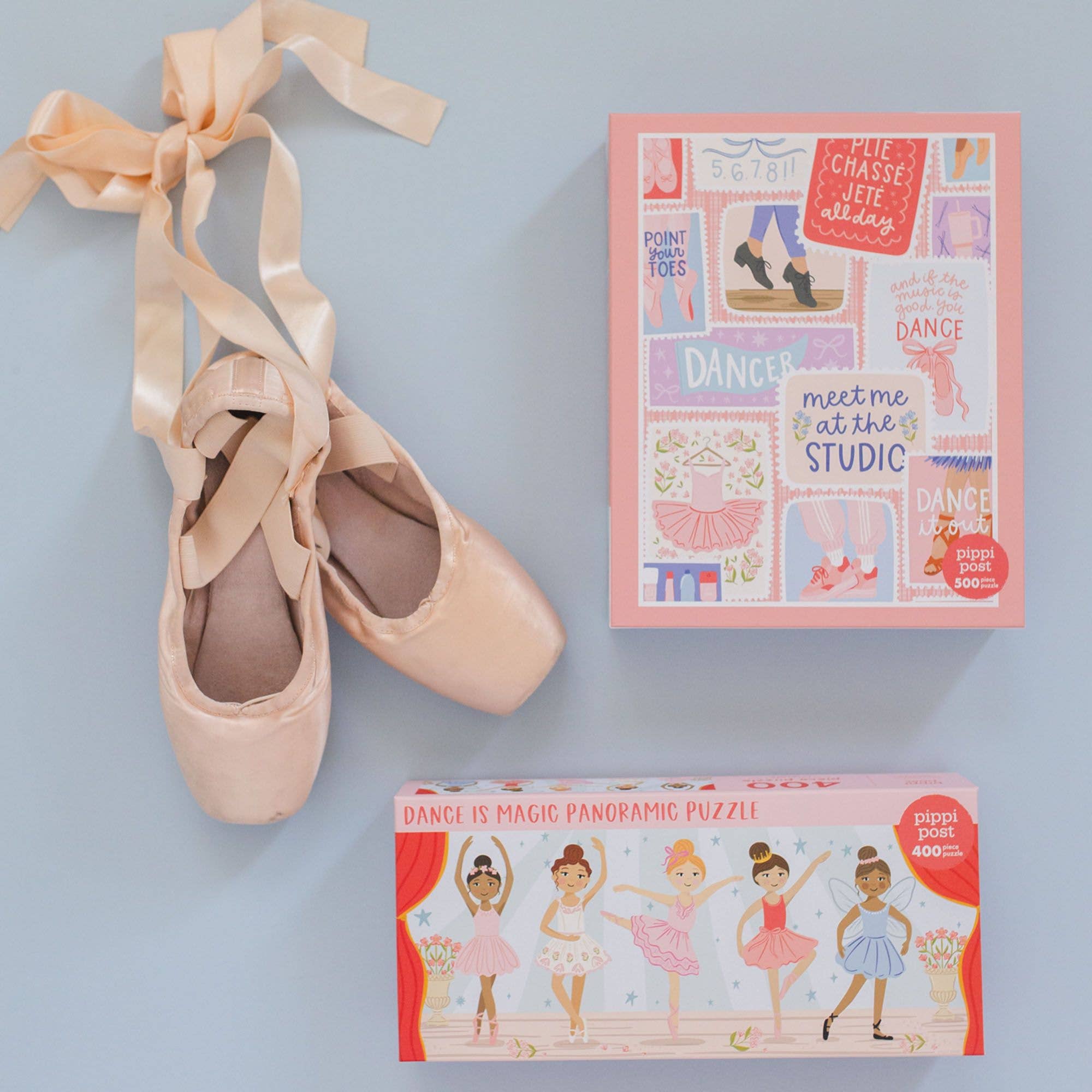 A pair of pale pink satin pointe shoes with long tied ribbons, shown beside two product boxes on a light blue background. The top box is pink with colorful ballet-themed illustrations and text, and the bottom box is red with ballerina artwork and the title 'Dance is Magic Panoramic Puzzle'.
