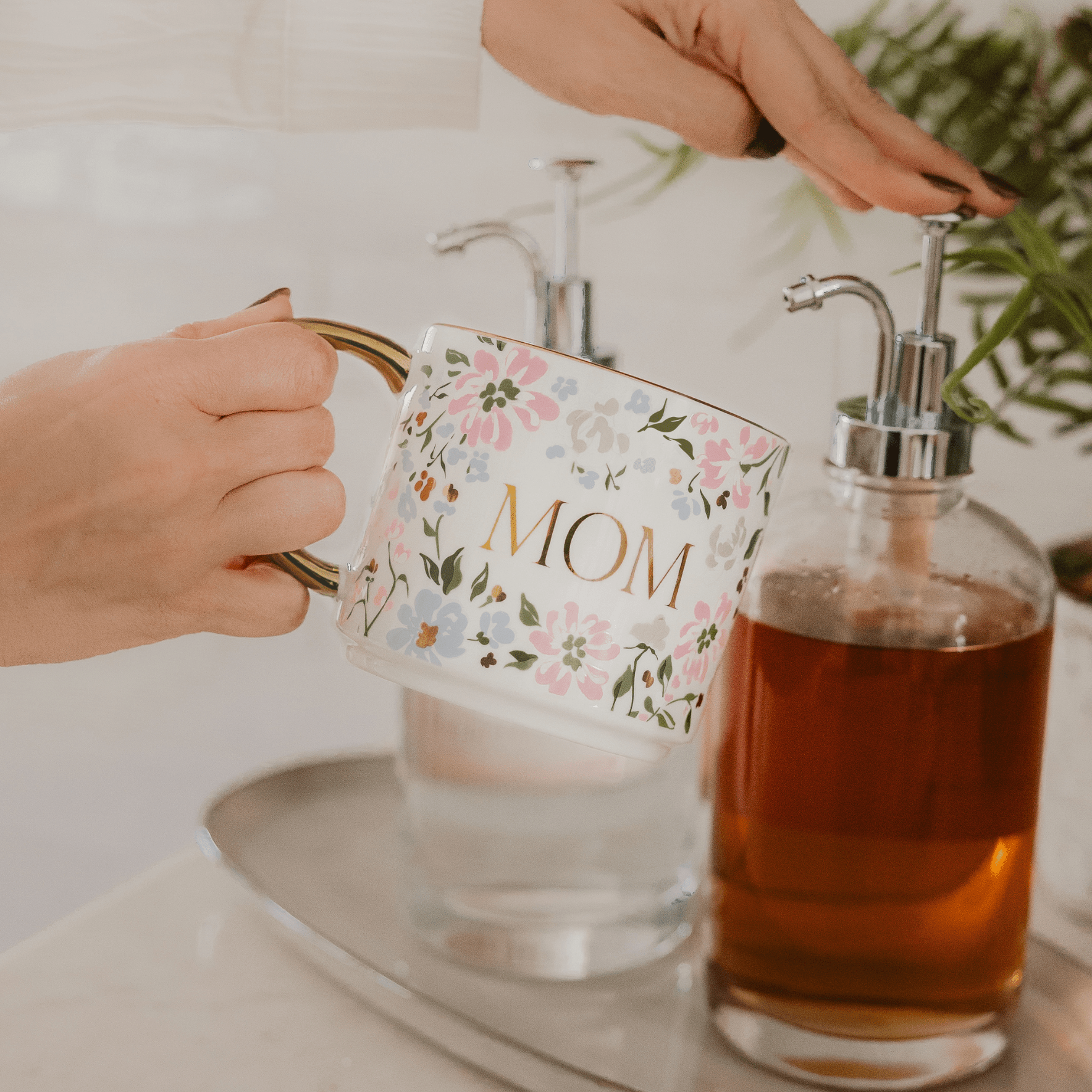 Person holding a floral mug labeled 'Mom' with a bottle of liquid soap on a counter.