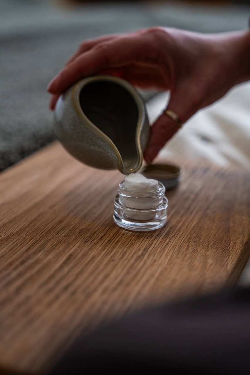 Hand pouring liquid from a ceramic pitcher into a glass on a wooden surface.