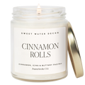 Sweet Water Decor Cinnamon Rolls scented candle in a clear glass jar with white wax, a lit wick, and a gold lid. Features a minimalist cream label with black lettering. Scent notes of cinnamon, icing, and buttery pastry. Made in the USA.