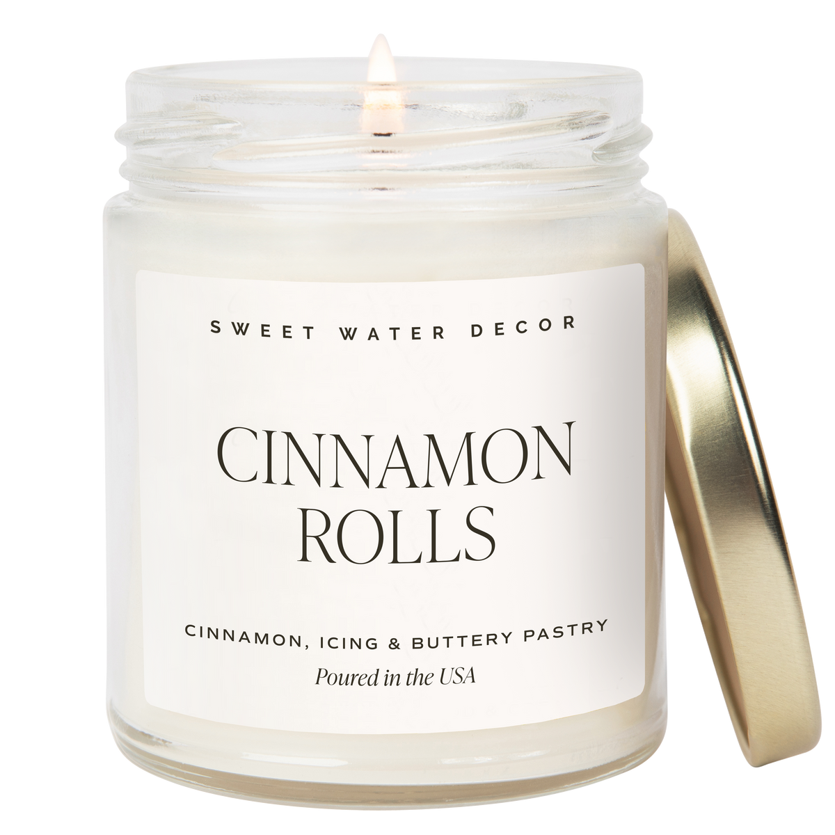 Sweet Water Decor Cinnamon Rolls scented candle in a clear glass jar with white wax, a lit wick, and a gold lid. Features a minimalist cream label with black lettering. Scent notes of cinnamon, icing, and buttery pastry. Made in the USA.