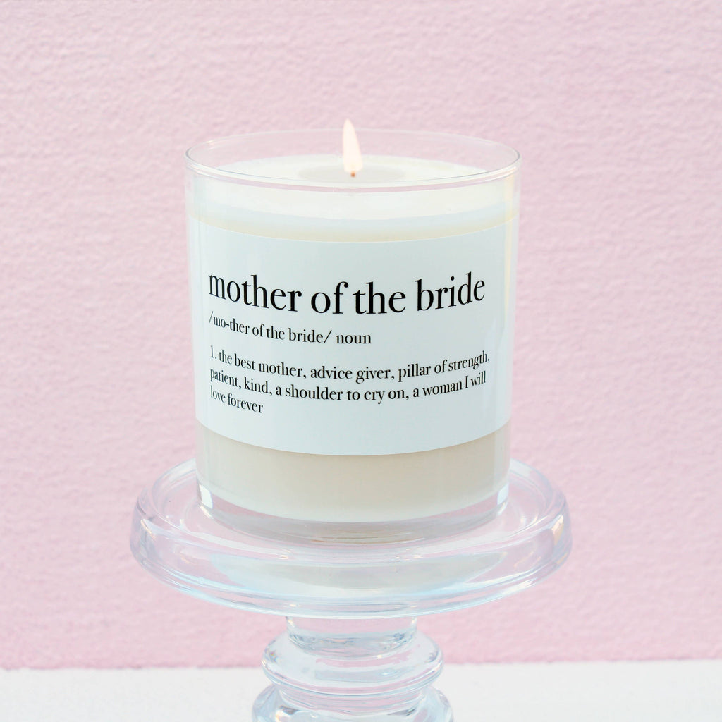 Mother of the Bride Definition Soy Candle in a white glass jar with a sentimental label, displayed on a clear pedestal against a soft pink background.
