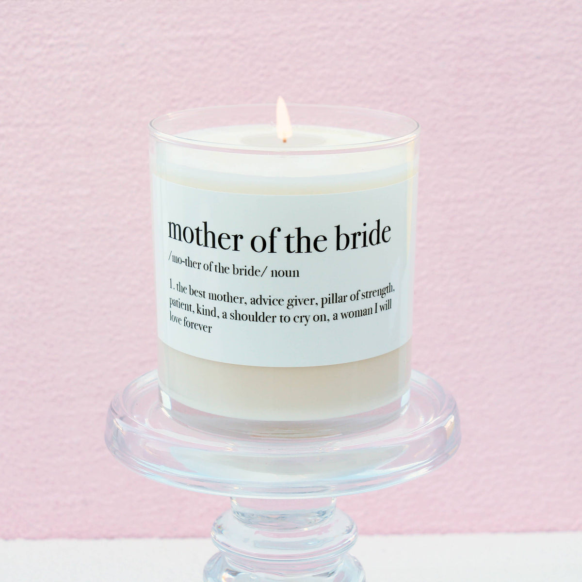 Mother of the Bride Definition Soy Candle in a white glass jar with a sentimental label, displayed on a clear pedestal against a soft pink background.
