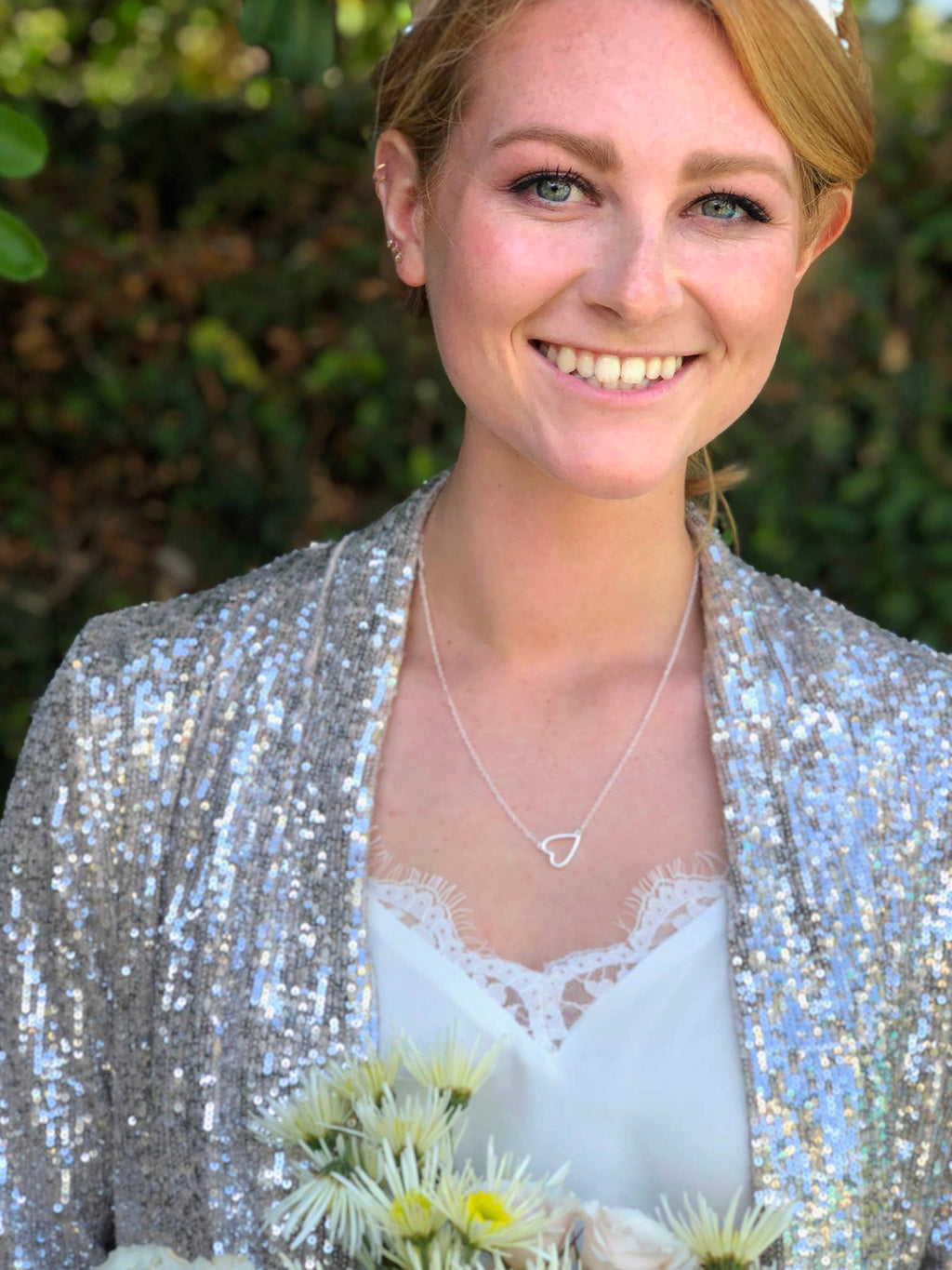 Woman wearing a silver sequin jacket and heart-shaped necklace, holding flowers with a blurred green background