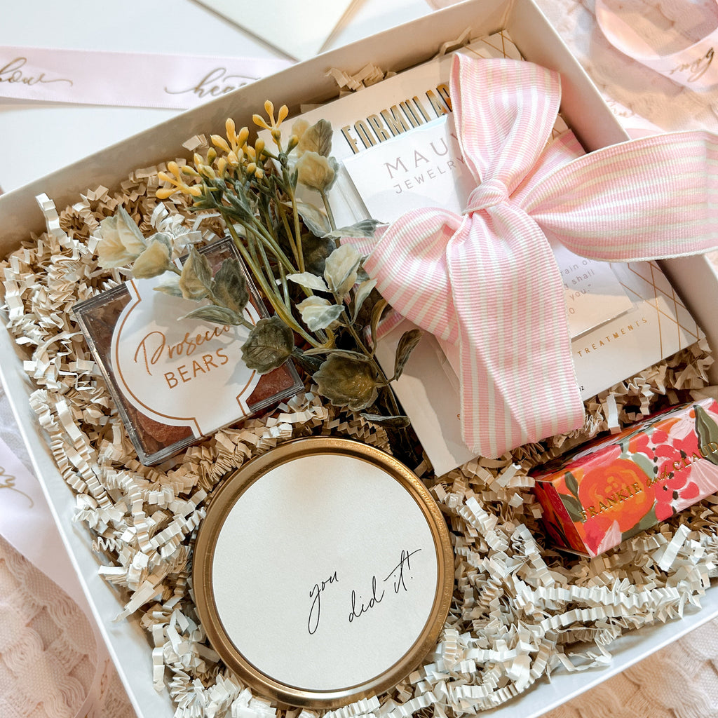 Gift box with decorative items including a small plant, a round container, and a pink ribbon on a light surface.