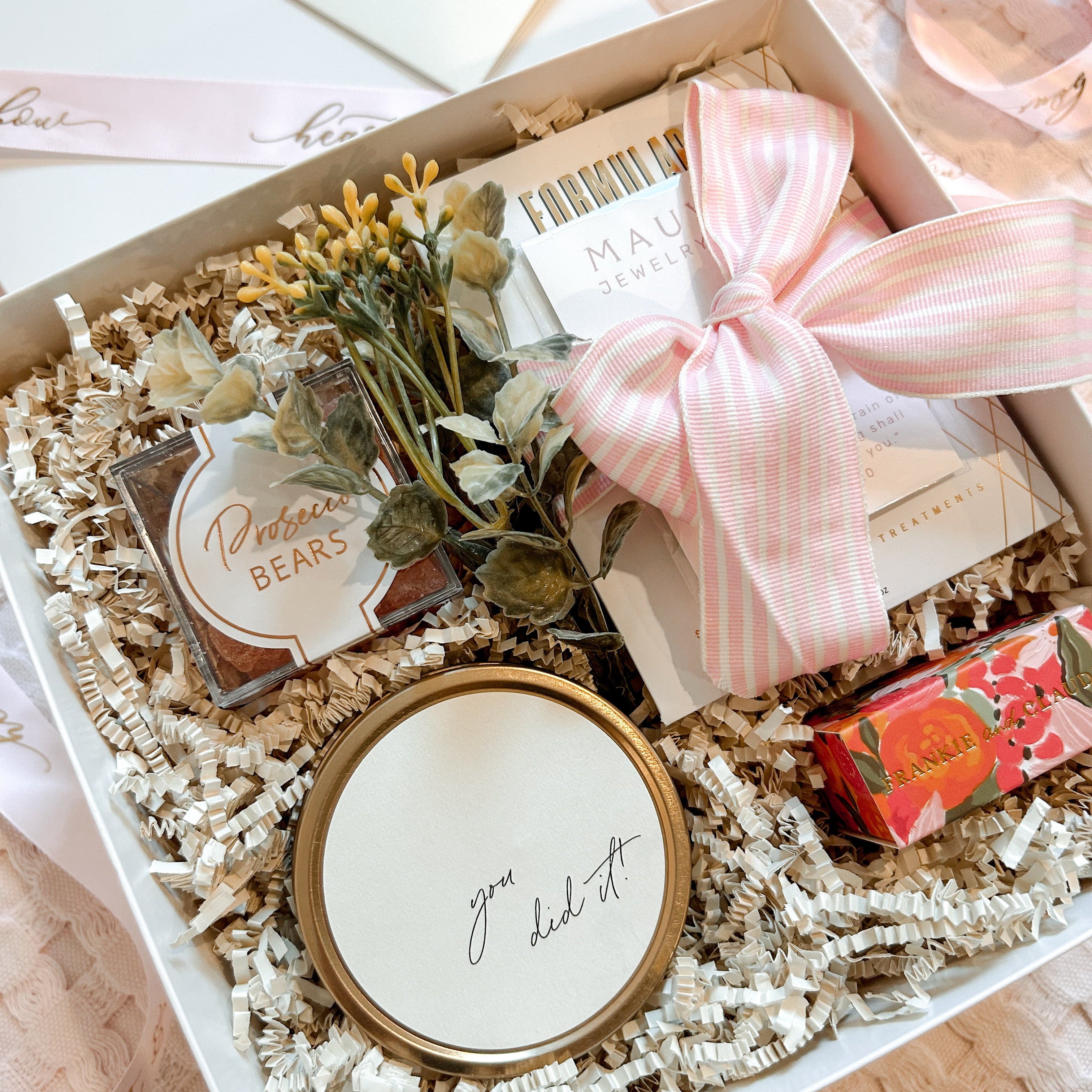 Gift box with decorative items including a small plant, a round container, and a pink ribbon on a light surface.
