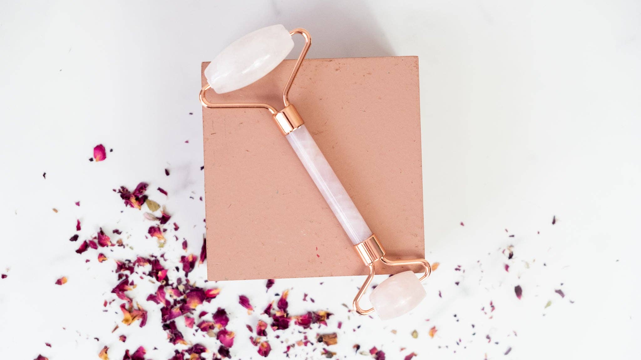 Rose gold and white facial roller on a pink block with scattered rose petals on a light background
