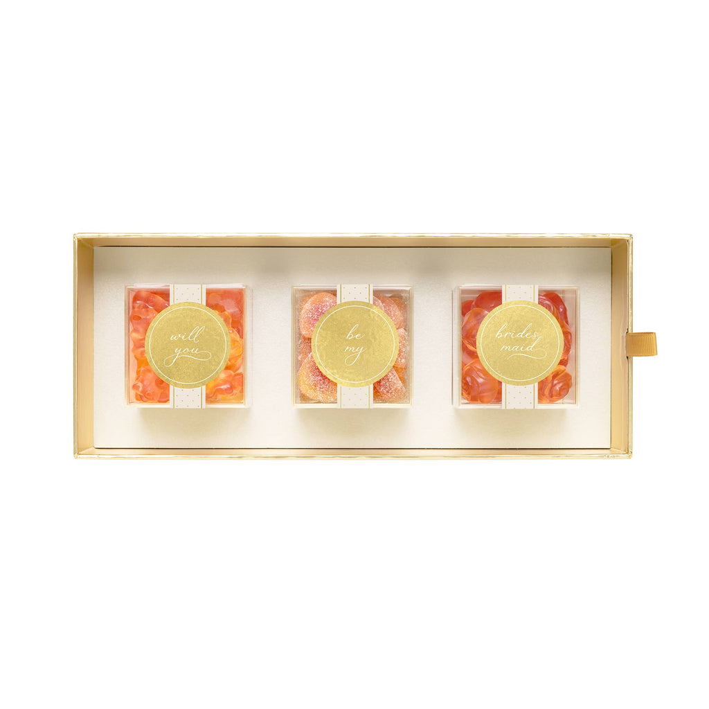 A rectangular gift box containing three small clear jars filled with orange-red candies, each topped with a round gold foil seal with elegant script text, set against a plain white background.