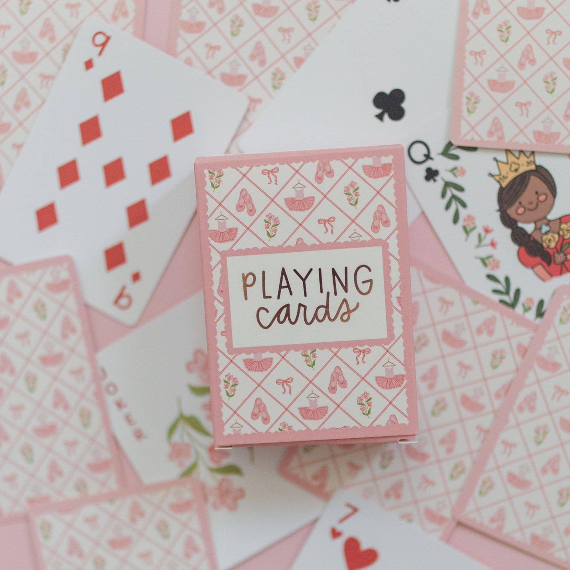 A pink and white playing cards box with a ballet theme, surrounded by scattered playing cards on a light pink background.