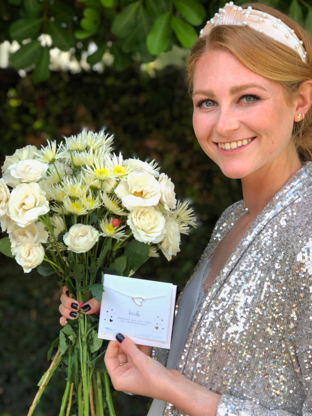 Woman in a silver sequin outfit holding flowers and a card with a green leafy background