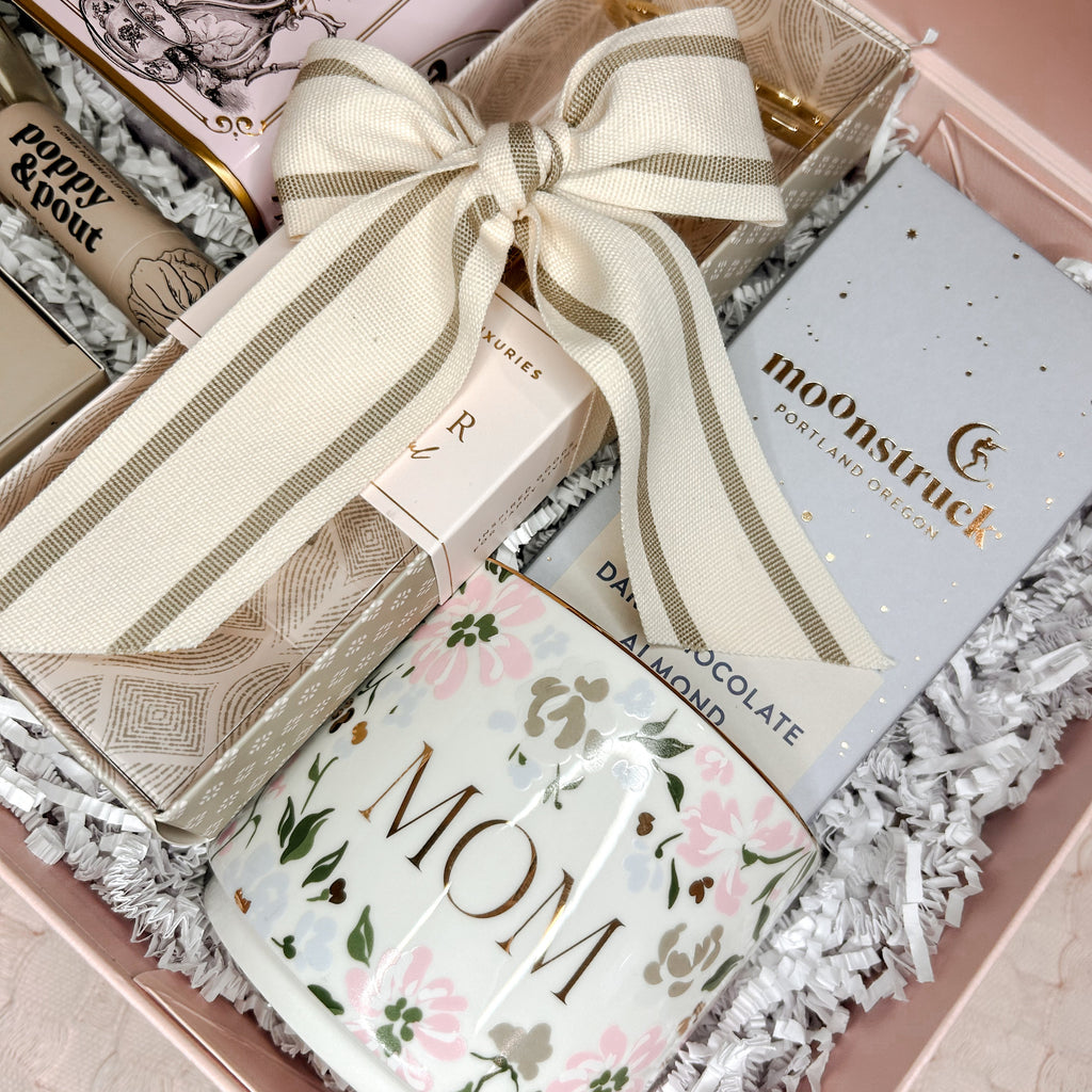 Gift box with tea packages, chocolates, and a 'Mom' card on a soft pink background.