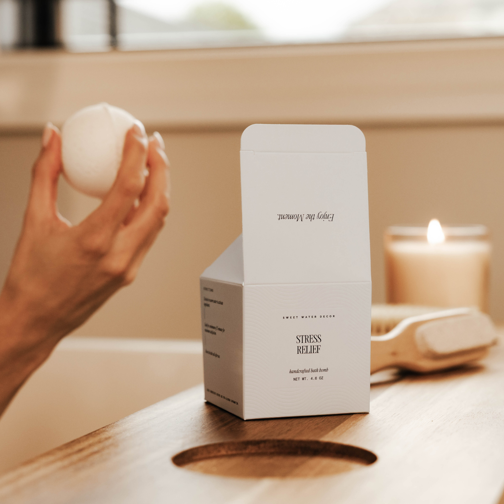 Hand holding a white bath bomb next to a box labeled 'Stress Relief' on a wooden surface with a blurred background.