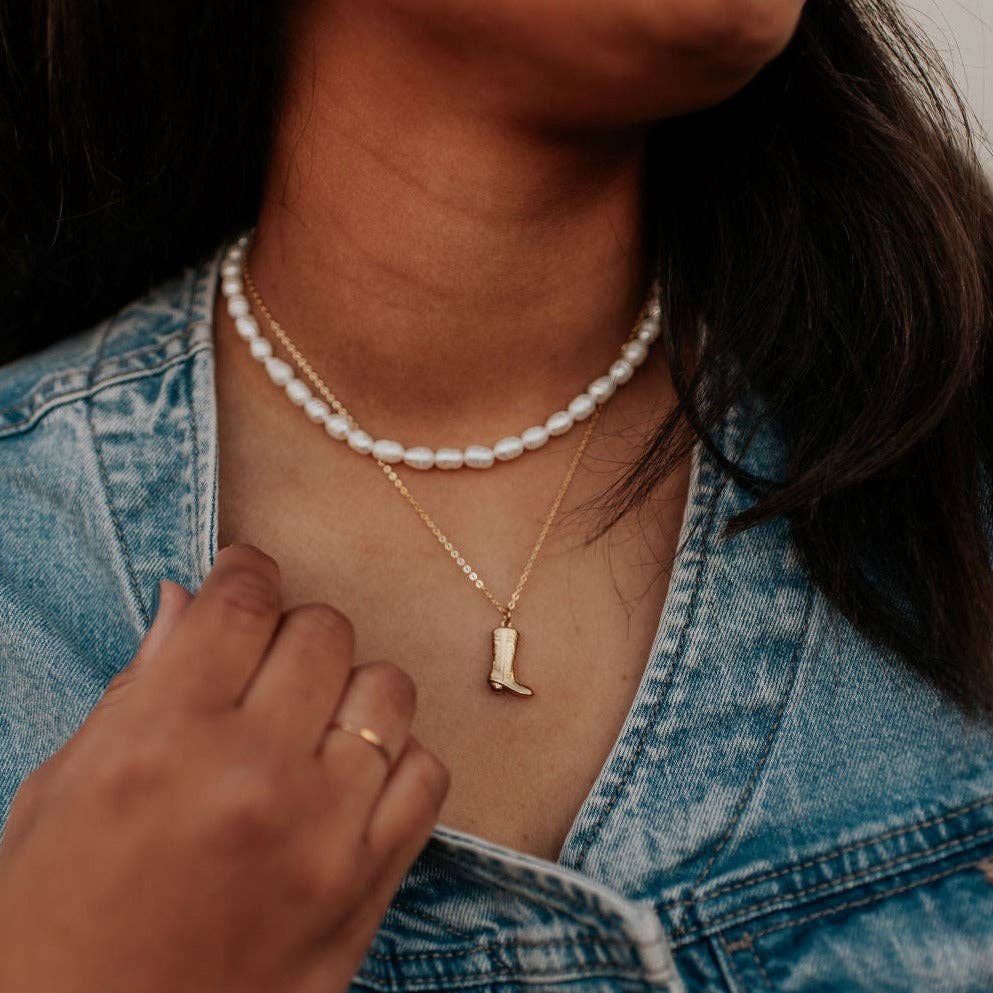Person wearing a pearl necklace and a gold necklace with a boot pendant, with a hand touching the pearl necklace.