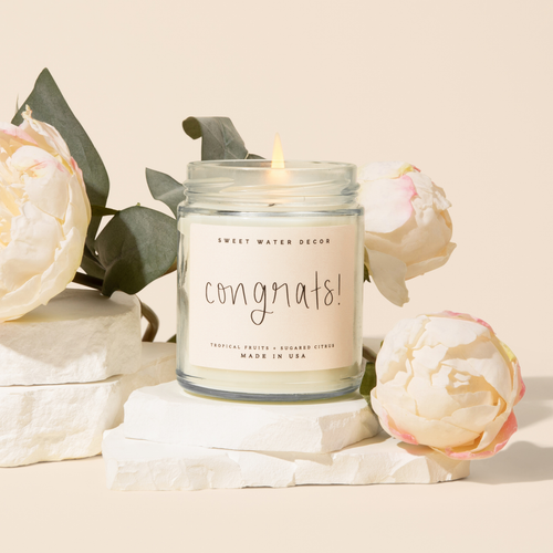 Candle in a jar labeled 'congrats!' surrounded by flowers on a light background