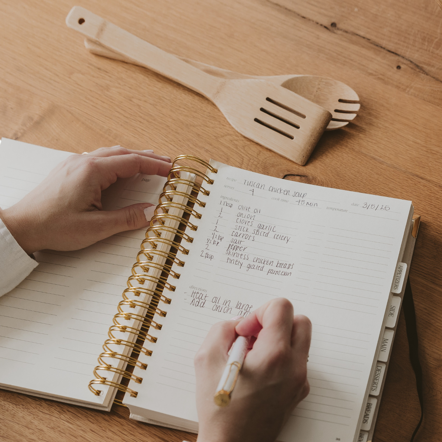 Person writing in a spiral-bound notebook on a wooden table with cooking utensils.