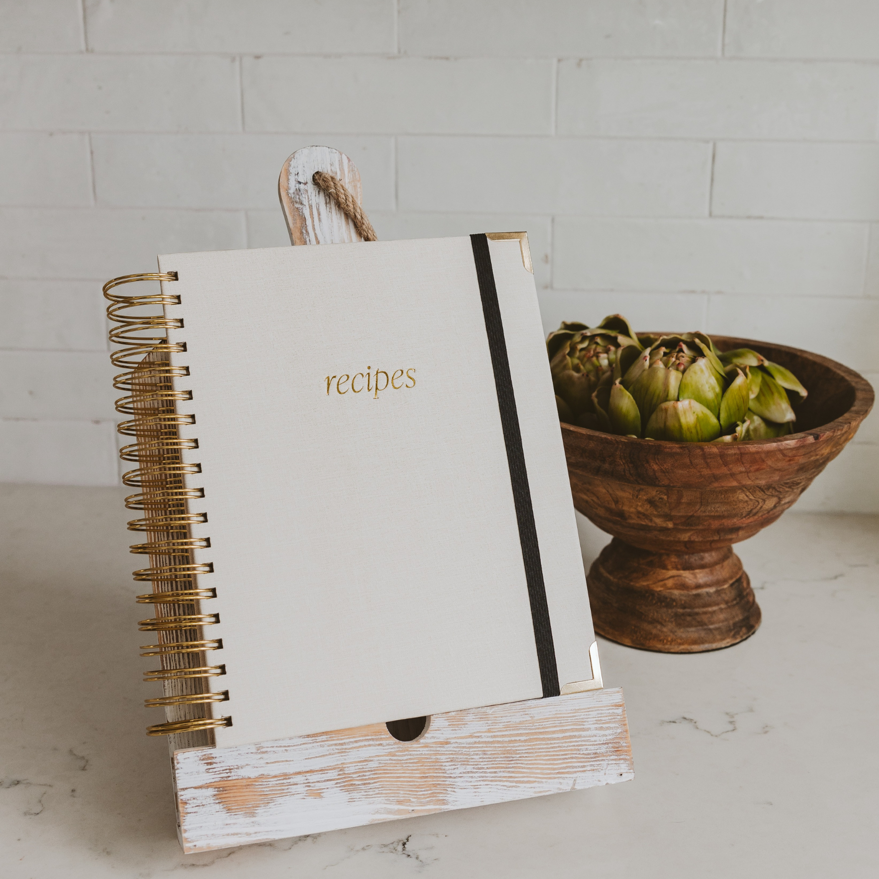 Recipe book with a wooden stand next to a bowl of artichokes on a marble surface.