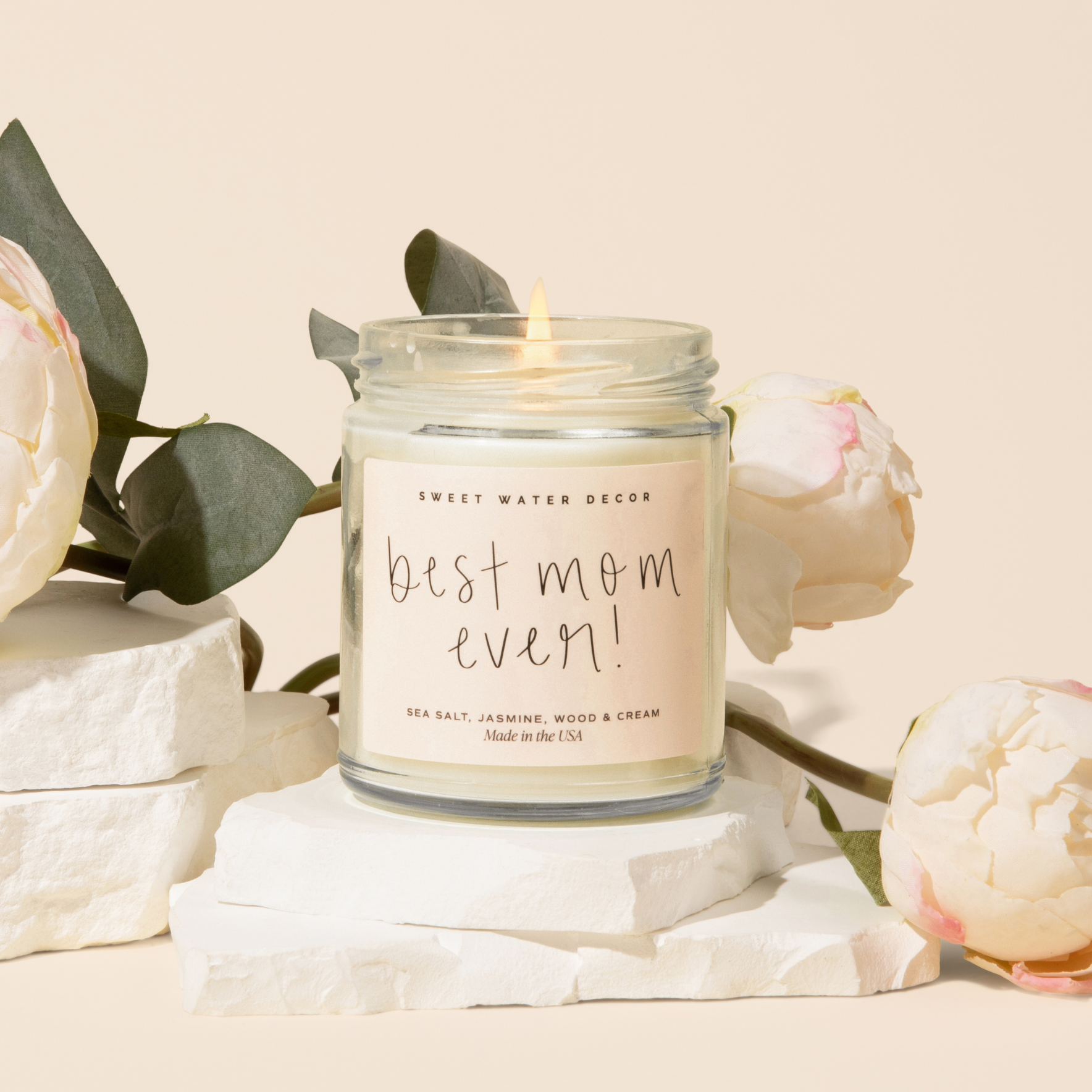 Candle in a jar labeled 'best mom ever' surrounded by flowers on a light background