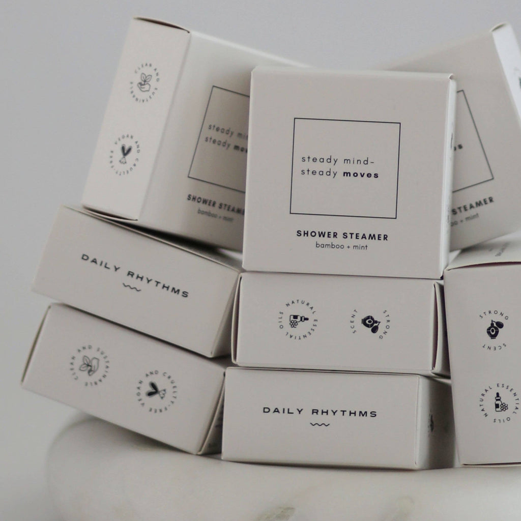 A stack of white rectangular boxes for shower steamers, with the front box reading 'steady mind, steady moves' and 'SHOWER STEAMER' beneath it. Several boxes show the brand name 'DAILY RHYTHMS'.