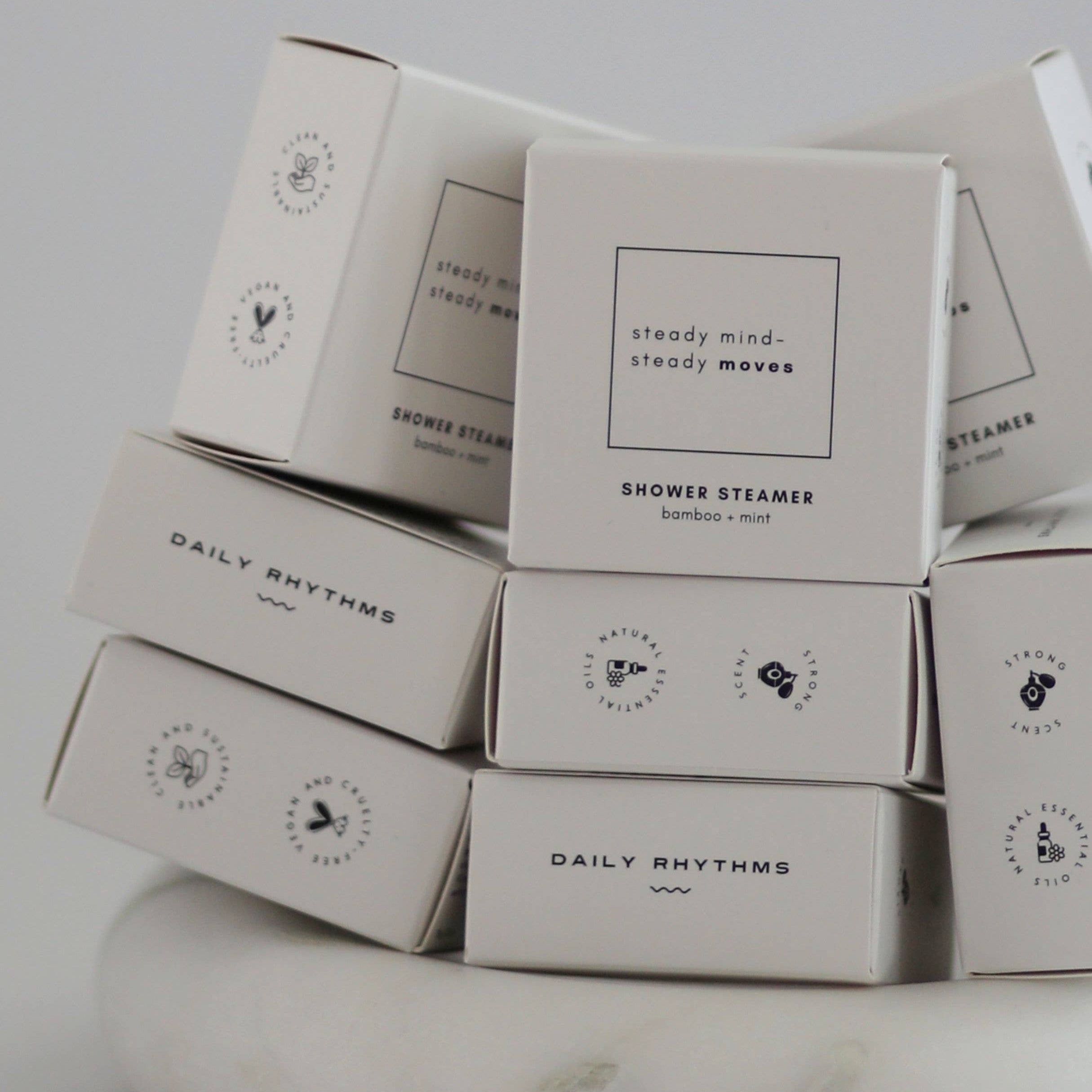 A stack of white rectangular boxes for shower steamers, with the front box reading 'steady mind, steady moves' and 'SHOWER STEAMER' beneath it. Several boxes show the brand name 'DAILY RHYTHMS'.