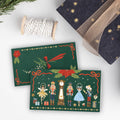 Two Christmas-themed greeting cards displayed on a white surface, surrounded by holiday props including a spool of white twine, a dark navy gift box with gold stars and ribbon, and evergreen branches. The cards feature festive green backgrounds with nutcracker illustrations and red poinsettias.