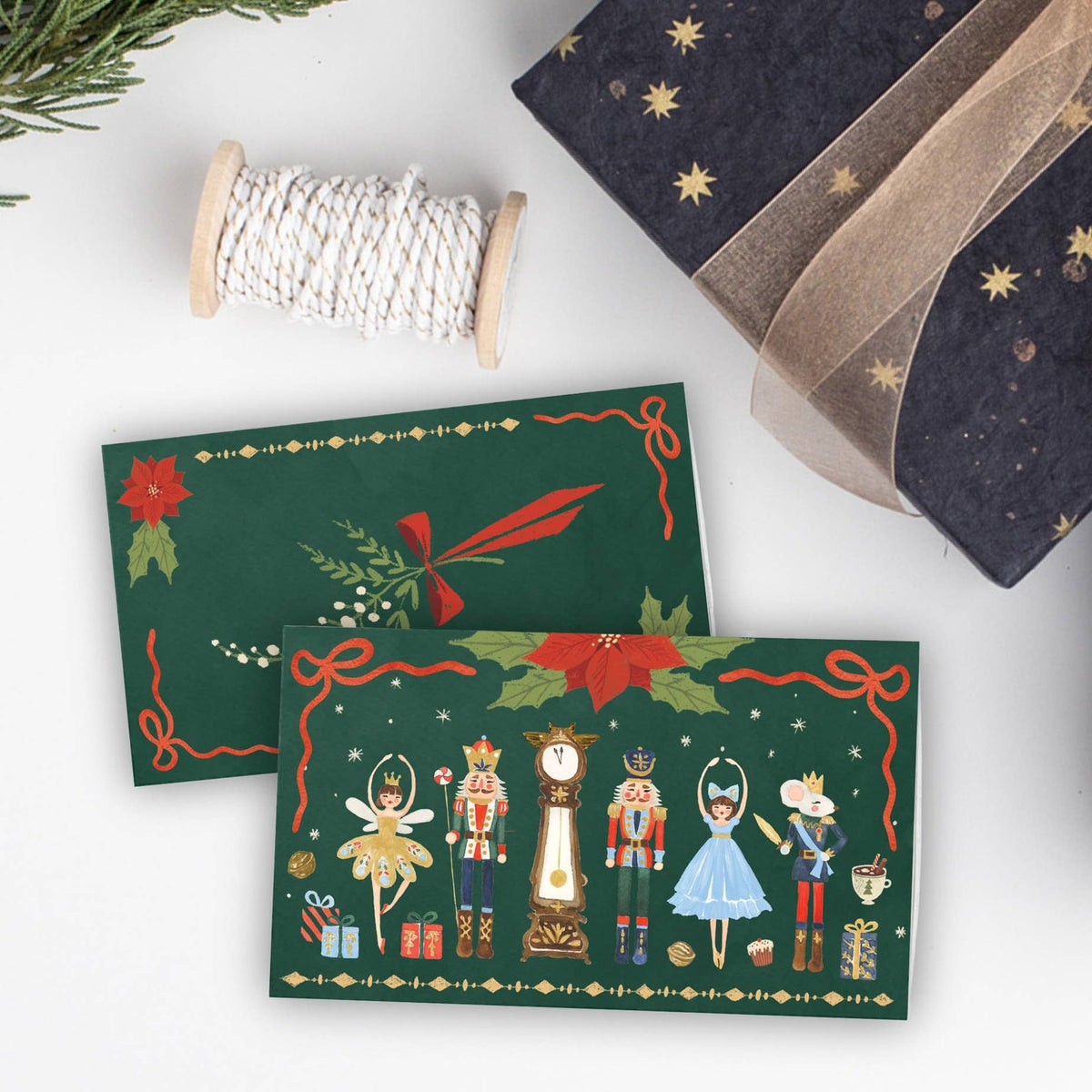 Two Christmas-themed greeting cards displayed on a white surface, surrounded by holiday props including a spool of white twine, a dark navy gift box with gold stars and ribbon, and evergreen branches. The cards feature festive green backgrounds with nutcracker illustrations and red poinsettias.