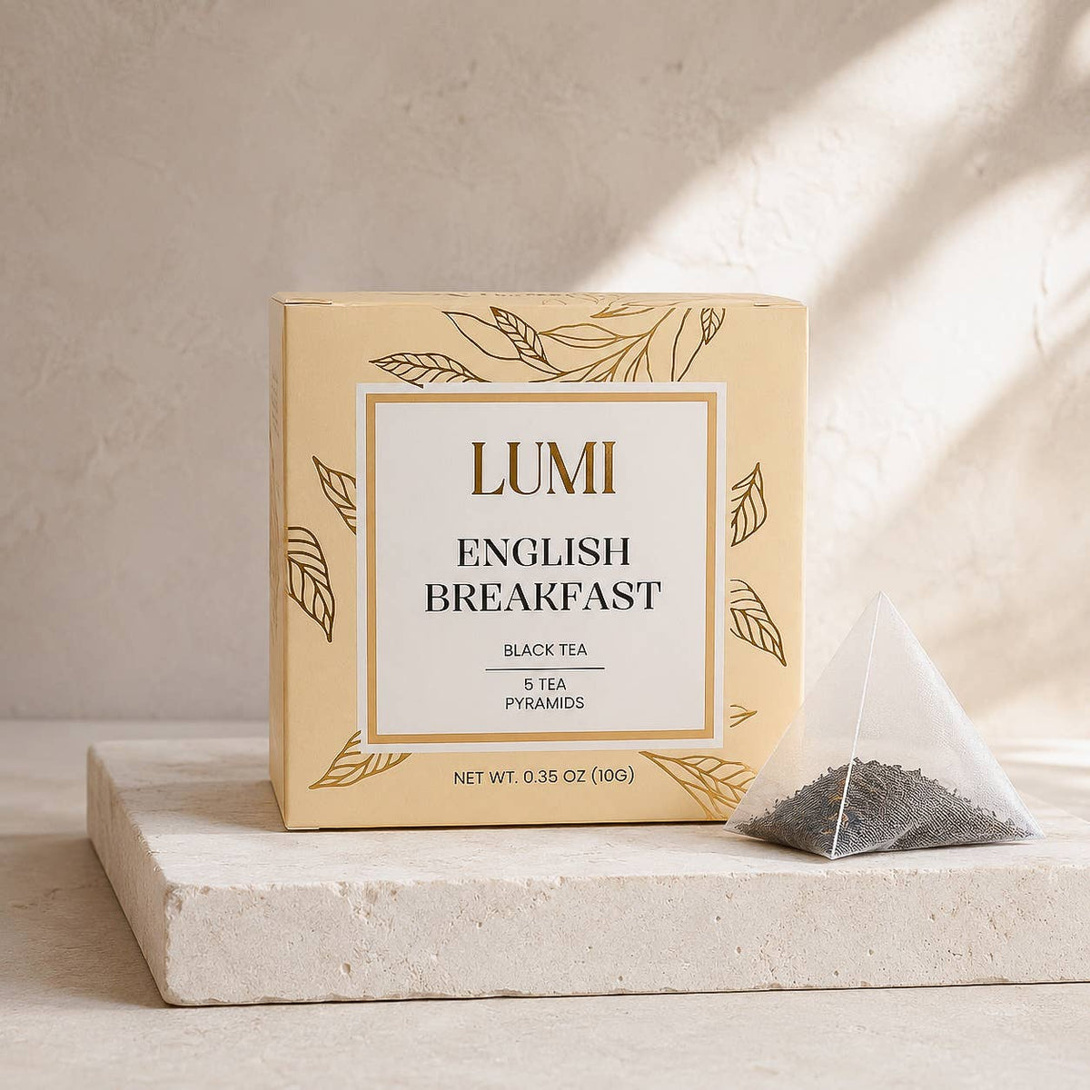 A box of LUMI English Breakfast black tea sits on a light stone pedestal with a single pyramid tea bag beside it. The packaging is cream with gold botanical line art, and the tea bag contains dark tea leaves.