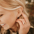 Close-up of a woman wearing multiple earrings and rings, with a blurred natural background.