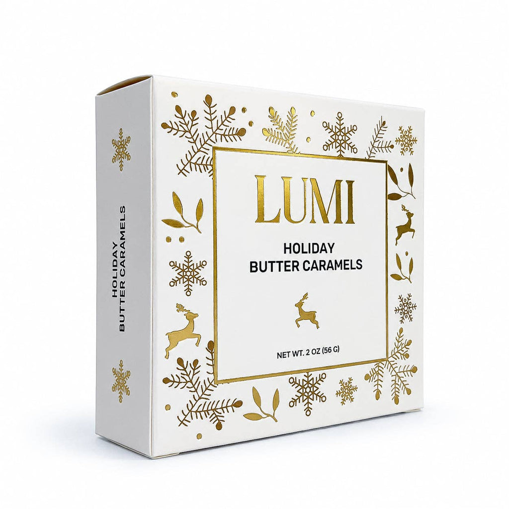 A box of Lumi Holiday Butter Caramels, featuring a white packaging design decorated with gold foil snowflakes, leaves, and deer, on a clean white background.