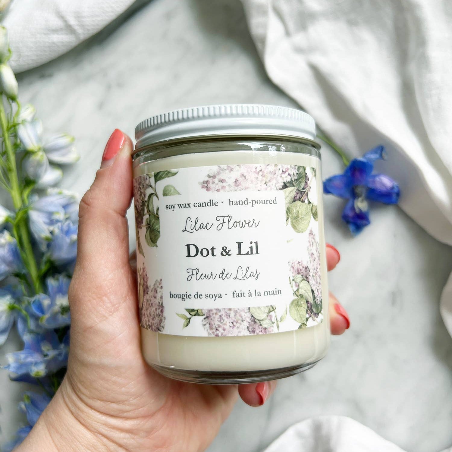 Hand holding a candle labeled 'Dot & Lil' with flowers in the background