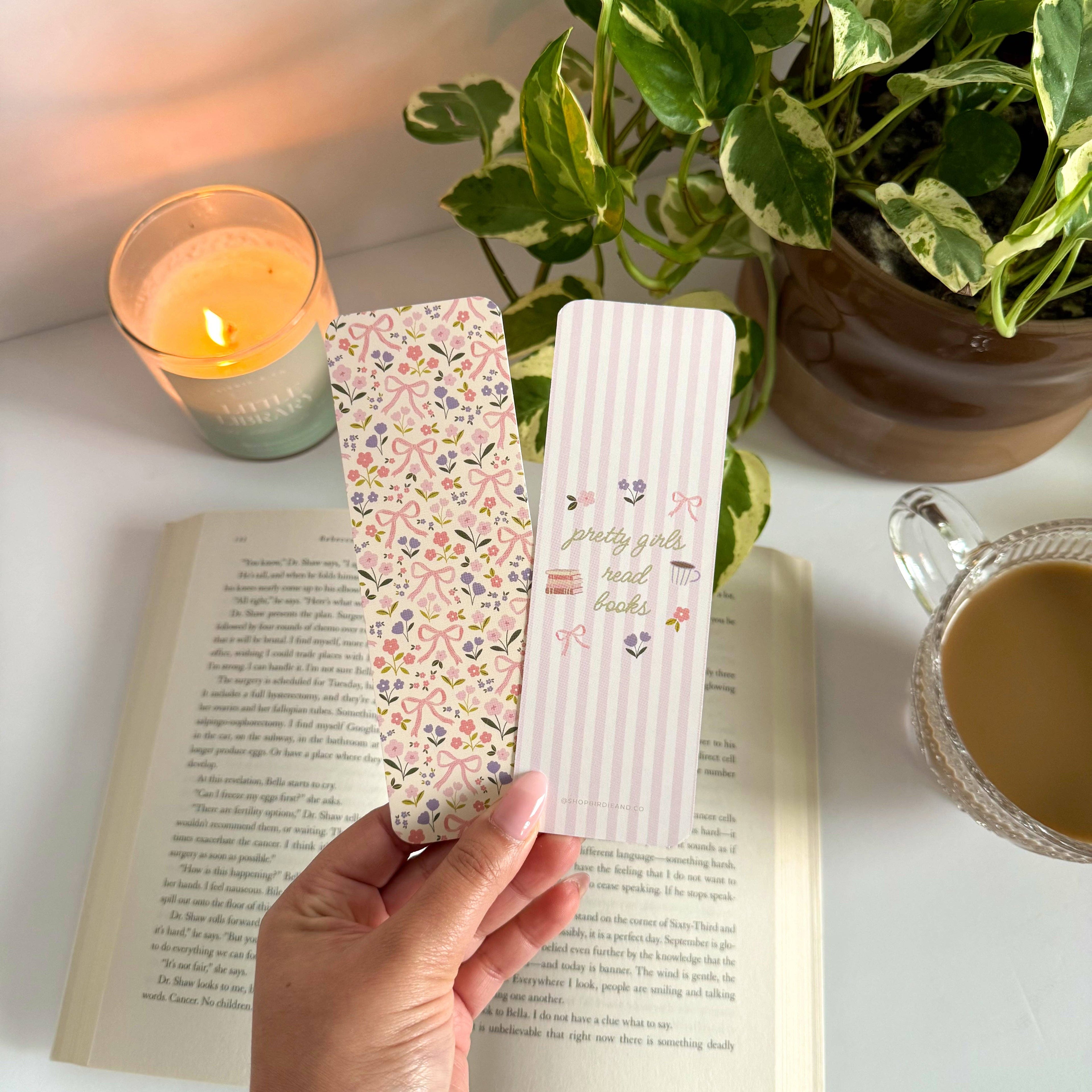 Two bookmarks with decorative designs held over an open book, with a candle and plant in the background.
