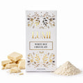 A white rectangular box of Lumi White Hot Chocolate with gold foil botanical illustrations and the brand name 'LUMI' in gold, shown on a white background with broken pale chocolate pieces on the left and a small pile of hot chocolate powder on the right.